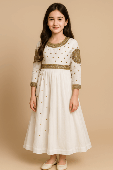 A young girl stands wearing a long, cream-colored A-line dress with three-quarter sleeves, embellished with small details and gold trim around the neckline, waist, and cuffs.