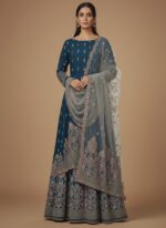 Unstitched deep teal (blue-green) formal suit fabric with elaborate gold, pink, and red traditional embroidery patterns on the lower half, and a separate light grey-green dupatta piece with scalloped cutwork and embroidery.