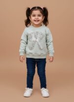 Toddler girl smiling in a mint green crewneck sweater with a sequin 'R', dark blue jeans, and white sneakers.