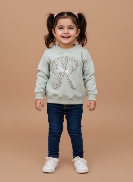Toddler girl smiling in a mint green crewneck sweater with a sequin 'R', dark blue jeans, and white sneakers.