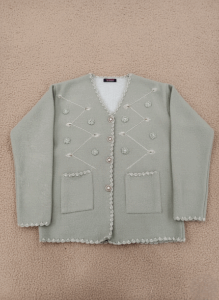 Women's sage green V-neck embellished button-front cardigan sweater with scallop trim and front pockets on a light brown background.
