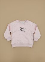 A light pink toddler sweater with small silver embellishments and a black "miu miu" logo in the center, laid flat against a beige background.