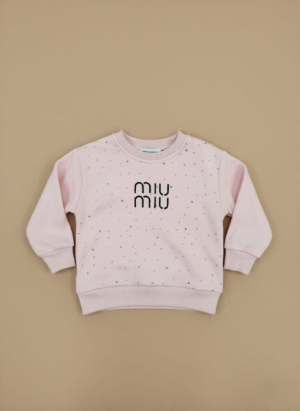 A light pink toddler sweater with small silver embellishments and a black "miu miu" logo in the center, laid flat against a beige background.