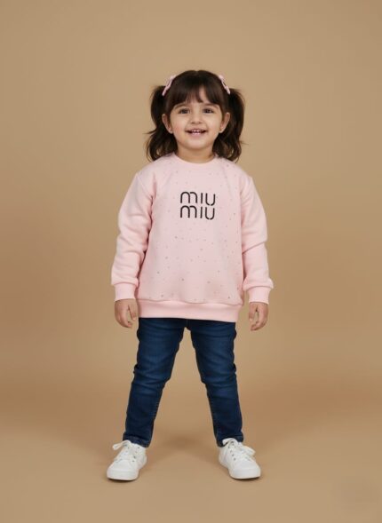 A smiling toddler girl wearing a light pink sweater with dark blue jeans and white sneakers, standing against a beige background. The sweater has "miu miu" written on it.