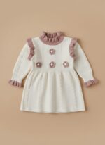 Cream colored baby girl knit sweater dress with pink ruffled collar, cuffs, and floral appliques, laid flat on a neutral background.