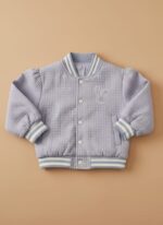 A happy toddler wearing a purple quilted bomber jacket, dark blue jeans, and white sneakers, standing against a beige background. The toddler quilted bomber jacket features ribbed cuffs and an embroidered design.
