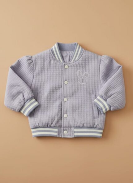 A happy toddler wearing a purple quilted bomber jacket, dark blue jeans, and white sneakers, standing against a beige background. The toddler quilted bomber jacket features ribbed cuffs and an embroidered design.