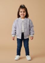 A happy toddler wearing a purple quilted bomber jacket, dark blue jeans, and white sneakers, standing against a beige background. The toddler quilted bomber jacket features ribbed cuffs and an embroidered design.