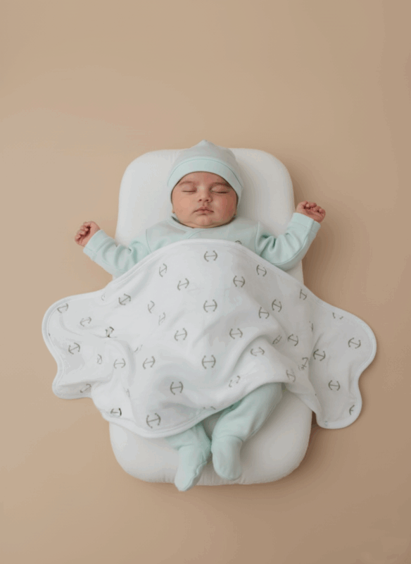 A sleeping baby dressed in a mint green long-sleeve bodysuit, pants, and hat, covered with a white baby swaddle blanket adorned with a grey anchor pattern.