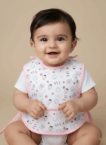 A smiling baby wearing a pink-trimmed cotton baby bib with a pattern of characters and stars, sitting comfortably.