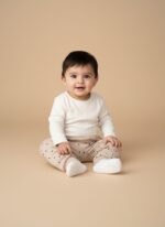 A smiling baby wearing a white long-sleeve top and beige baby jogger pants with black polka dots.