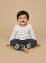 A smiling baby wearing a white long-sleeve top and dark grey ribbed baby leggings with bow and pom-pom details on the ankles.