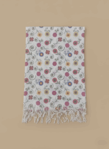 White floral print shawl with colorful flowers, green swirls, and white fringe laid flat on a beige background.