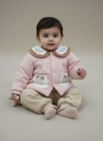 A happy baby sitting and looking at the camera, dressed in a pink long-sleeve sweater featuring a white Peter Pan collar with floral embroidery and a brown ruffled edge, two decorative pockets with plaid bows, and beige corduroy pants. This is the Baby Peter Pan Collar Sweater Set.