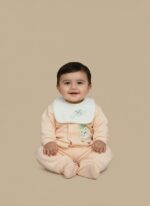 Baby wearing a light peach sweater with a white bib, featuring a cute animal appliqué on the sweater and embroidered clouds on the bib, paired with matching peach pants.