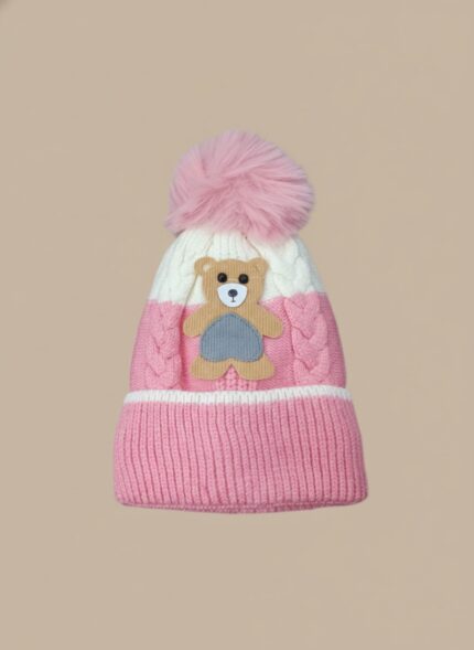 A knitted baby hat in shades of grey and white with a pink bear appliqué on the front and a large, fluffy grey faux fur pompom on top.
