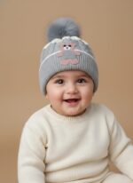 A knitted baby hat in shades of grey and white with a pink bear appliqué on the front and a large, fluffy grey faux fur pompom on top.