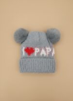 A knitted baby hat in shades of grey and white with a pink bear appliqué on the front and a large, fluffy grey faux fur pompom on top.