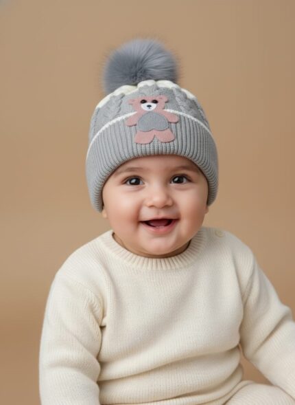 A knitted baby hat in shades of grey and white with a pink bear appliqué on the front and a large, fluffy grey faux fur pompom on top.