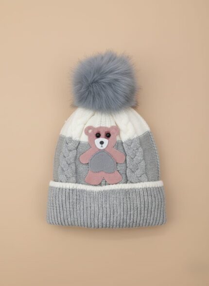 A knitted baby hat in shades of grey and white with a pink bear appliqué on the front and a large, fluffy grey faux fur pompom on top.