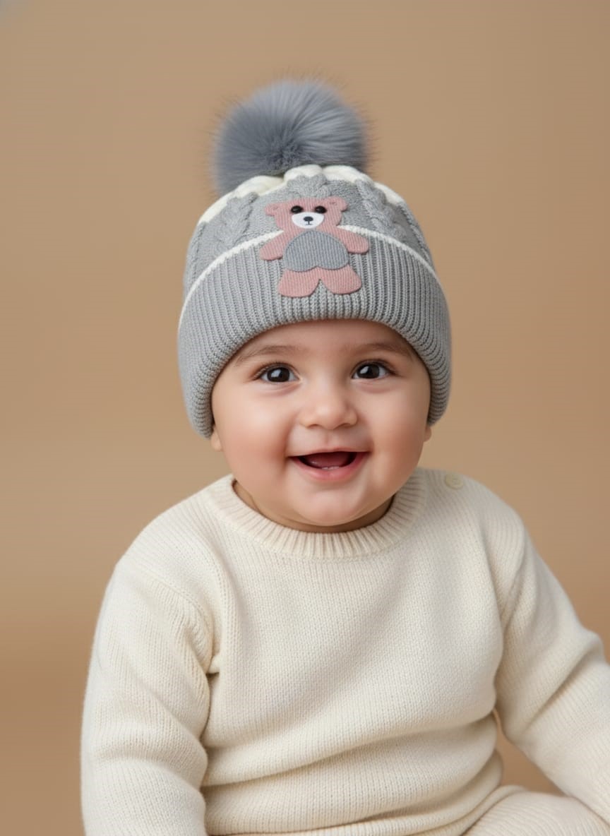 Baby Bear Pom Hat with Faux Fur Pompom A knitted baby hat in shades of grey and white with a pink bear appliqué on the front and a large, fluffy grey faux fur pompom on top.