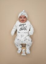 Smiling baby wearing a white, black-polka-dot "Welcome to the World" four-piece starter outfit set, including a knotted hat, bib, long-sleeve onesie, and footed pants.