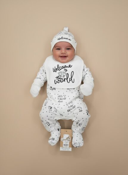 Smiling baby wearing a white, black-polka-dot "Welcome to the World" four-piece starter outfit set, including a knotted hat, bib, long-sleeve onesie, and footed pants.