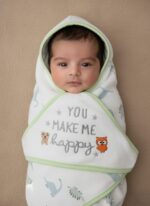 A newborn baby swaddled in a white blanket with green trim, featuring "YOU MAKE ME HAPPY" embroidered text, a teddy bear, an owl, and dinosaur patterns.