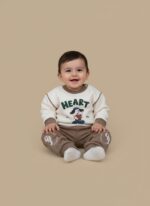 A happy baby sitting and smiling, dressed in a cream long-sleeve sweater with a "HEART" text and a dog graphic holding a heart, paired with brown jogger pants with white graphics on the knees. This is the Baby Heart Dog Sweater Set.