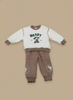 A happy baby sitting and smiling, dressed in a cream long-sleeve sweater with a "HEART" text and a dog graphic holding a heart, paired with brown jogger pants with white graphics on the knees. This is the Baby Heart Dog Sweater Set.