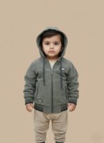 Close-up of a smiling Pakistani toddler wearing an olive green zip-up hooded jacket on a light brown background. Kids casual wear.