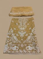 A highly detailed, unstitched Mustard Chiffon Embroidered Dress material featuring heavy, intricate white threadwork and subtle gold accents, laid flat with a matching embroidered dupatta/pallu piece above it. Includes raw silk trouser material