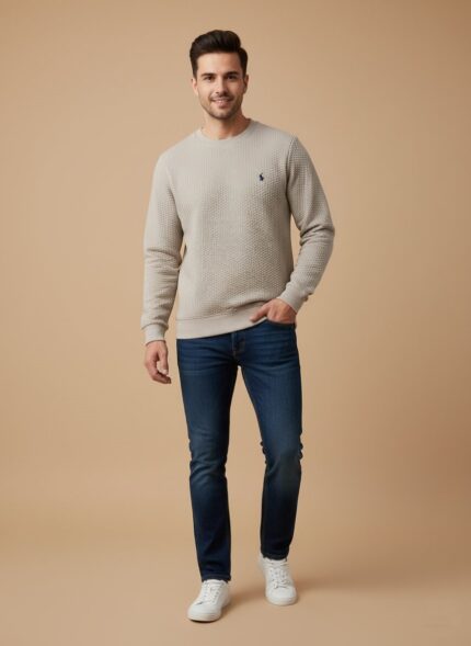 Full shot of a smiling man wearing a beige textured crewneck sweater, blue jeans, and white sneakers, posing casually.