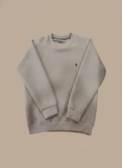 Full shot of a smiling man wearing a beige textured crewneck sweater, blue jeans, and white sneakers, posing casually.