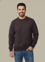 Smiling man wearing a dark charcoal quilted crewneck sweater with a small logo, paired with blue jeans, posing with hand in pocket.