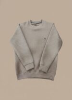 Image of a man wearing a beige waffle knit sweater with a crew neck and a small embroidered logo, dark blue jeans, and white sneakers, standing against a beige background.