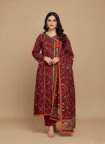 A woman wearing a vibrant maroon floral printed khaddar Angrakha style suit with a long-sleeved, flared kurta, matching maroon trousers, and a coordinating printed khaddar dupatta. The suit features colorful floral prints and orange lace trim.