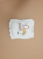 White baby pillow with a colorful car, palm tree, and sun illustration, set against a light brown background.