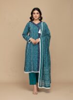 A woman wearing a vibrant teal printed khaddar Angrakha style suit with a long-sleeved shirt, matching trousers, and a coordinating printed khaddar dupatta. The suit features geometric prints, white lace trim, and a tassel.