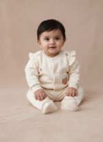 A happy baby sitting and smiling, dressed in a cream long-sleeve sweater featuring a bunny appliqué holding a strawberry, small strawberry embroideries, and ruffle shoulder details, paired with matching cream pants. This is the Baby Bunny & Strawberry Outfit.
