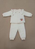 A happy baby sitting and smiling, dressed in a cream long-sleeve sweater featuring a bunny appliqué holding a strawberry, small strawberry embroideries, and ruffle shoulder details, paired with matching cream pants. This is the Baby Bunny & Strawberry Outfit.