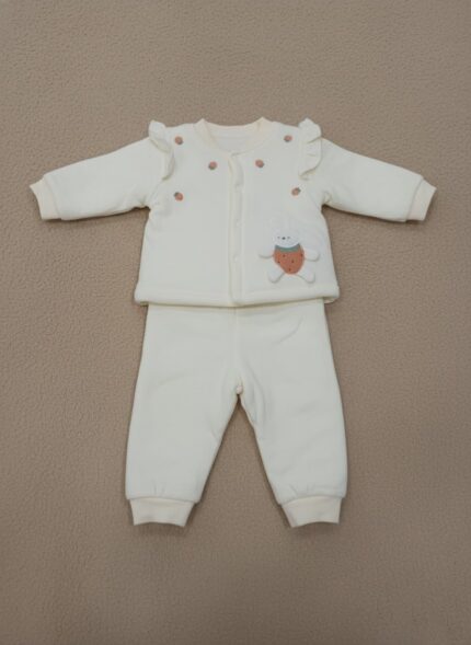 A happy baby sitting and smiling, dressed in a cream long-sleeve sweater featuring a bunny appliqué holding a strawberry, small strawberry embroideries, and ruffle shoulder details, paired with matching cream pants. This is the Baby Bunny & Strawberry Outfit.