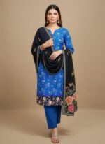 A woman wearing a royal blue suit set. The kurta has subtle floral prints and a colorful floral border at the hem. She is wearing matching blue trousers and a draped black shawl with large, colorful floral embroidery at the ends.