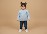 Smiling toddler girl in a light blue knit sweater with bunny appliques and bows, blue jeans, and white canvas shoes, standing on a beige background.