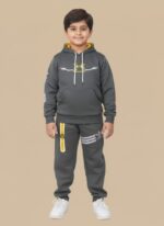 Happy boy in a grey athletic tracksuit with a yellow-lined hoodie and sports logos, matching joggers with stripes, and white sneakers, posing against a beige background.