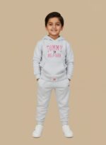 Full-length image of a smiling young boy wearing a light grey Tommy Hilfiger Boys Grey Tracksuit with a logo hooded sweatshirt and matching joggers, standing in white sneakers against a beige background.