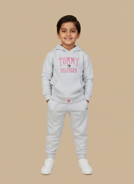 Full-length image of a smiling young boy wearing a light grey Tommy Hilfiger Boys Grey Tracksuit with a logo hooded sweatshirt and matching joggers, standing in white sneakers against a beige background.