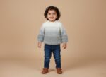 Happy toddler girl in a fluffy white and grey ombre sweater, blue jeans, and brown Mary Jane shoes, standing on a beige background.