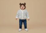Happy toddler girl in a fluffy white and grey striped sweater with buttons, blue jeans, and white canvas shoes, standing against a beige background.