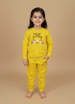 Full-length image of a smiling young girl wearing a bright yellow Girls Yellow Tracksuit set, consisting of a "Cute and Purrfect" cat-themed sweatshirt and matching joggers, standing barefoot against a beige background.